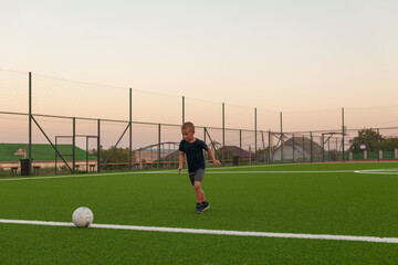 A boy dressed in a blue T-shirt and gray shorts on a football field covered with artificial green...