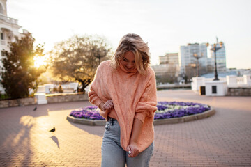 Fototapeta na wymiar Stylish girl in a peach sweater walks in the street of the city against the background of sunset sun and flowers. Portrait of smiling woman
