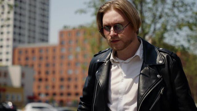 An angry man in a leather jacket walks through a city block.