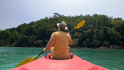 Young man with sunglasses and hat rows pink plastic canoe along sea against green hilly islands...