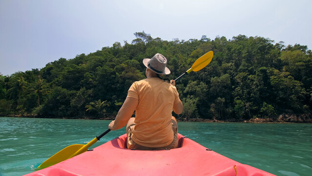 Young man with sunglasses and hat rows pink plastic canoe along sea against green hilly islands with wild jungles. Traveling to tropical countries. Strong guy is sailing on kayak in ocean, back view.