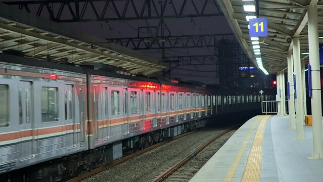 A commuter train full of passengers comes into station in Jakarta at night with a lit-up high-rise building in the background. It depicts the crowded train during work hours. 