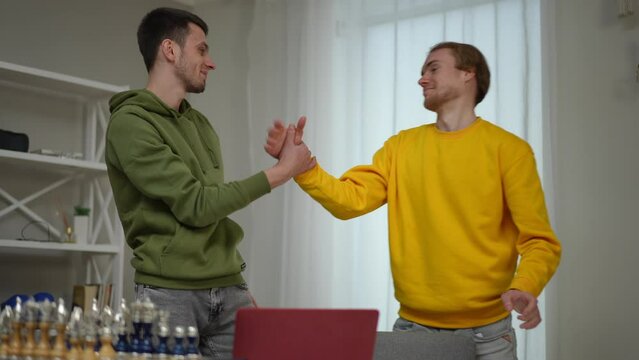Satisfied young Caucasian men surfing Internet on laptop gesturing high-five smiling. Positive successful freelancers rejoicing startup success indoors