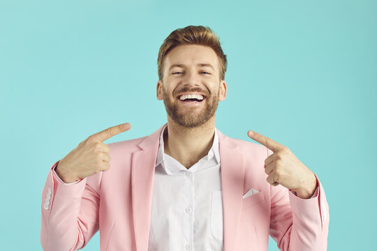 Happy, satisfied man in a pink suit and with a perfect smile points at his mouth, shows his even, straight teeth, and says that he likes the result of professional whitening and orthodontic correction