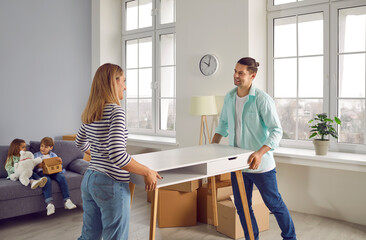 Happy family with two kids moving into new home. Young parents carrying table and unpacking cardboard boxes. Smiling little children playing on sofa in living room