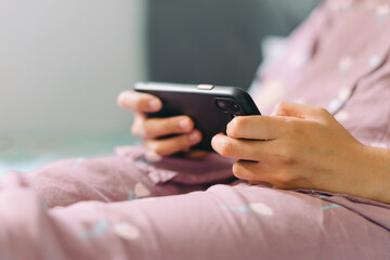 Close up woman hand in pajamas lies on a bed looking at a smartphone screen. Streaming watching leisure concept