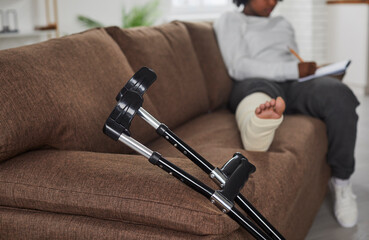 Close up of black elbow crutches by brown sofa, with African American woman with broken leg in cast...