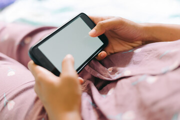Close up woman hand in pajamas lies on a bed looking at a smartphone with a white screen. Streaming leisure concept