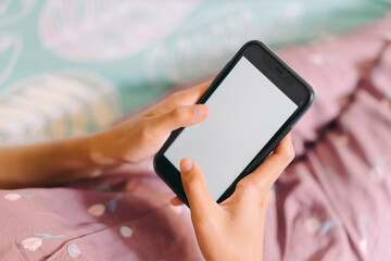 Close up woman hand in pajamas lies on a bed looking at a smartphone with a white screen. Streaming leisure concept