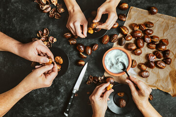 Close up of hands of three people peeling roasted chestnuts on black background. Lifestyle family meal concept. Dark low key photo. Top view.