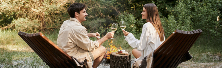 Side view of smiling couple holding wine near food and firewood during vacation, banner
