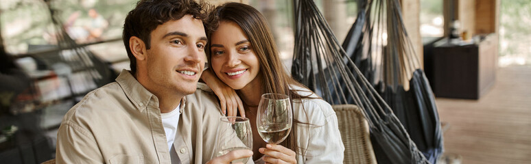 Smiling brunette woman hugging boyfriend and holding wine on terrace of vacation house, banner