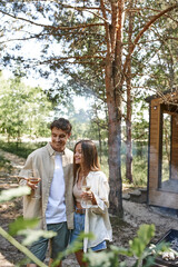 Naklejka na ściany i meble Joyful couple in casual clothes holding wine near barbecue and blurred vacation house at background