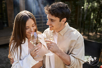 Cheerful man hugging girlfriend with wine near bbq and vacation house at background