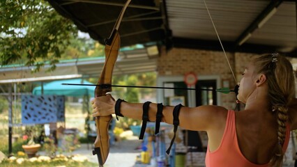 Fototapeta na wymiar Sport woman in shooting range with bow. Sportsman in shooting gallery aim an arrow to hit target. Outdoor archery training. Practice and training of archery in shooting range. Athlete keep wooden bow