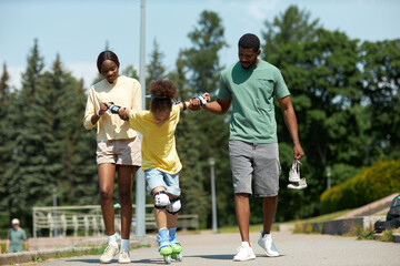 Parents holding hands with child and teaching her to roller skating in the park