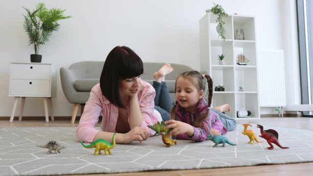 Beautiful lady in cozy outfit holding dinosaurs toy while talking with adorable preteen girl in modern apartment. Affectionate mother-daughter duo sharing mutual gaze while roleplaying animal world.