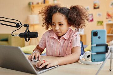 Cute serious schoolgirl looking at laptop screen while sitting by desk in front of computer and recording new podcast or making livestream