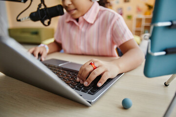 Focus on hand of schooolgirl typing on laptop keyboard while sitting by desk and recording podcast or making stream for online audience