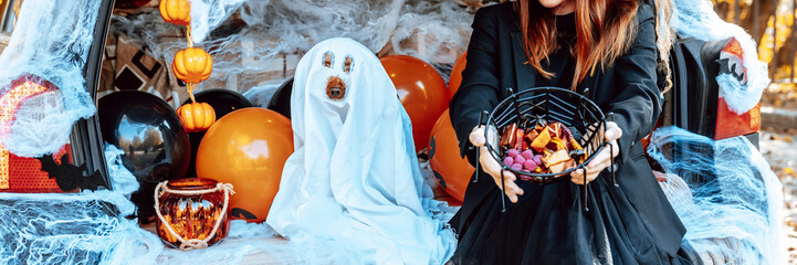 teenage girl in witch costume and hat and cute poodle dog in ghost costume sits in trunk car decorated for Halloween with web, orange balloons and pumpkins, outdoor creative activity concept in autumn