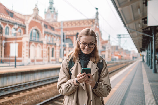 Tourist 30s female stands on the platform of railway station near passenger train car with mobile phone in hands. High quality photo
