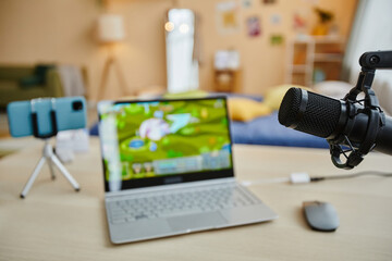 Focus on microphone hanging in front of camera against desk with video game on screen of laptop, smartphone on tripod and desktop mouse
