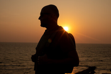 Portrait of traveler in sunglasses walking with backpack near sea on seashore at sunset, standing...