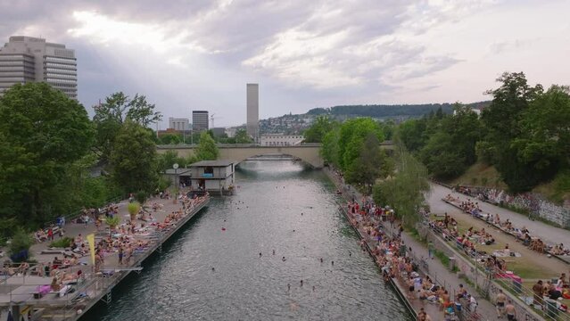 Forwards fly above public swimming area in river in city. People relaxing in water or on bank on warm day. Zurich, Switzerland