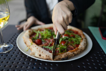Closeup of caucasian woman client hand cutting pizza with tomatoes, prosciutto and arugula with a round knife on a plate background in the restaurant or cafe. Italian fast food. Dry wine glass on a