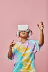 Modern schoolgirl in t-shirt and VR headset reaching to virtual object while playing three dimension video game against pink background