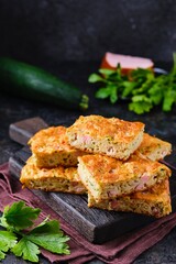 Sliced snack cake with zucchini, ham and cheese on a wooden board on a dark concrete background. Recipes for zucchini.