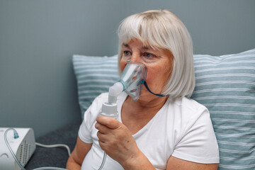 Sick upset caucasian senior woman with an inhaler. Unhealthy female doing inhalation at home, she use nebulizer and inhaler for the treatment, sitting on the couch at home, need treatment.  - obrazy, fototapety, plakaty