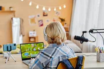 Rear view of blond schoolboy typing on laptop keyboard while sitting by desk in front of camera in home environment and playing video game