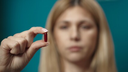 Obraz premium Close up shot of blonde woman model holding a red pill in her fingers, showing it to the camera