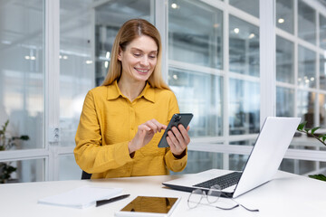 Mature experienced business woman inside office at workplace, employee sitting at desk smiling holding phone in hand, boss using application on smartphone, browsing internet pages.