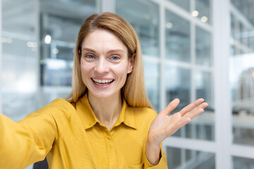 Online video call, businesswoman inside office at workplace looking at smartphone camera using video call app talking with friends and smiling joyfully.