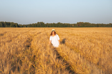 A girl in a hat walks through a wheat field at sunset