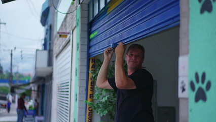 Man closing storefront garage in city sidewalk. Person slides storefront in street