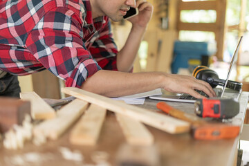 Asian man owner a small furniture business is recheck order online. carpenter male is adjust wood to the desired size. architect, designer, Built-in, professional wood, craftsman, workshop.