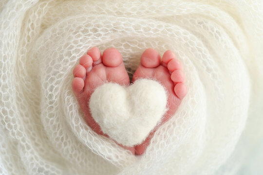 Knitted white heart in the legs of a baby. The tiny foot of a newborn baby. Soft feet of a new born in a white wool blanket. Close up of toes, heels and feet of a newborn. Macro photography