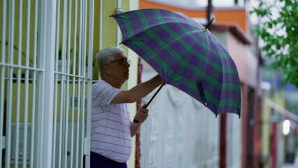 Older man arriving home while raining, opens gate and closes umbrella. Senior walking in street...
