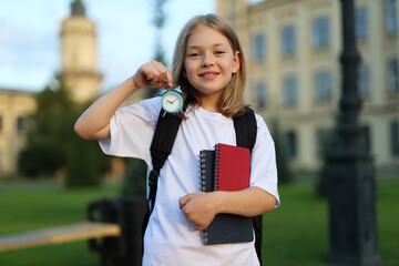 A cute schoolgirl in a white shirt and a backpack holds a notebook and an alarm clock.