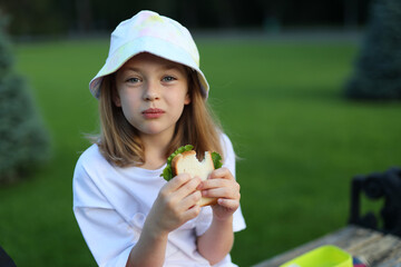 A girl with a backpack and a sandwich sitting at a picnic. She is going back to school and having lunch in the garden.