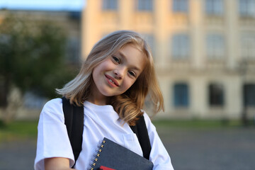 A young schoolgirl with a backpack and a notebook walks in front of a school building on a sunny day.