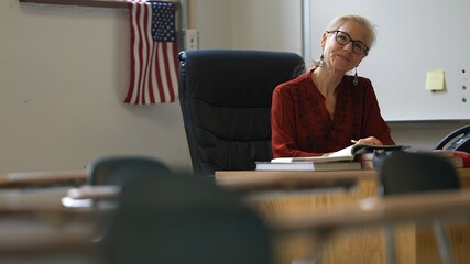 Portrait of happy smiling woman teacher at school classroom desk gives big smile. US American flag in the background.