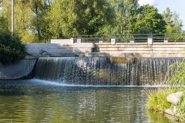 A small beautiful waterfall on a quiet calm river in a city park and a concrete footbridge over it