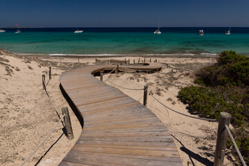 Wooden path to the sandy beach on the island of Formentera and view of the emerald water of the Mediterranean Sea and yachts