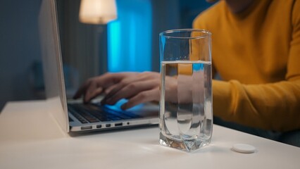 Effervescent tablet and a glass of water on the table close up. In the background, a man's hands are typing on a laptop. Cure for a cold, pain or hangover. Home medicine concept.