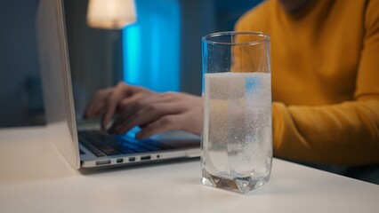 An effervescent tablet dissolves in a glass of water on a table close up. In the background, a man's hands are typing on a laptop. Cure for a cold, pain or hangover. Home medicine concept.