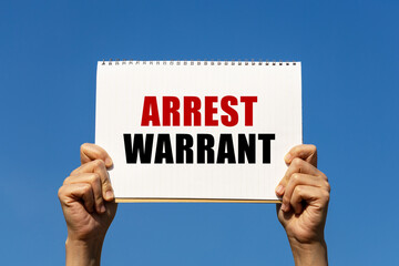 Arrest warrant text on notebook paper held by 2 hands with isolated blue sky background. This message can be used as business concept about arrest warrant.